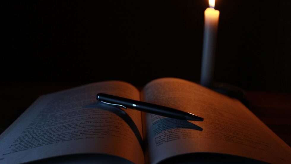 Libro aperto e penna su una scrivania illuminata da una candela, atmosfera raccolta per la lettura.