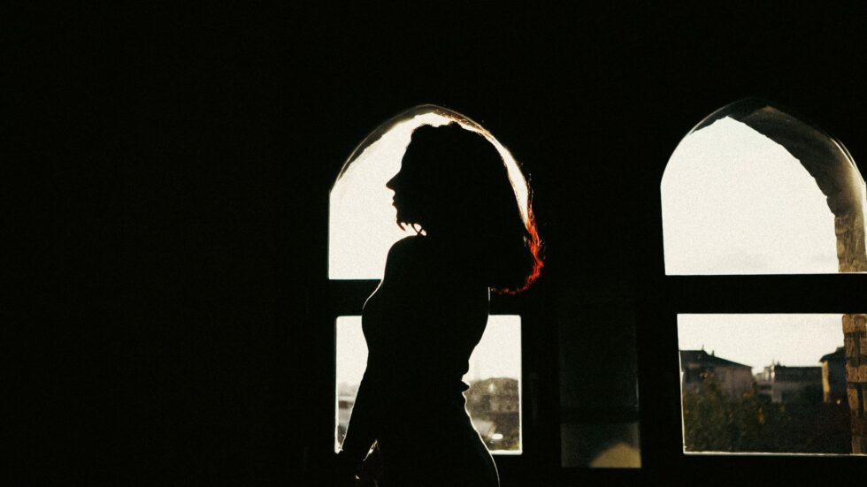 Silhouette di una donna in piedi accanto a una finestra ad arco, con un'ombra drammatica sulla parete